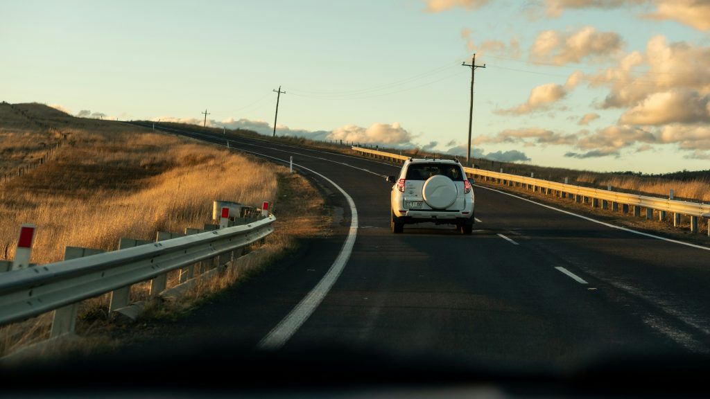 Een witte SUV rijdt een bochtige plattelandsweg op tussen goudkleurige velden onder een deels bewolkte lucht.