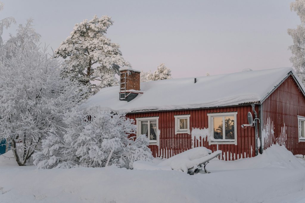 A red wooden house covered in snow, surrounded by snow‑laden trees under a soft pastel winter sky.