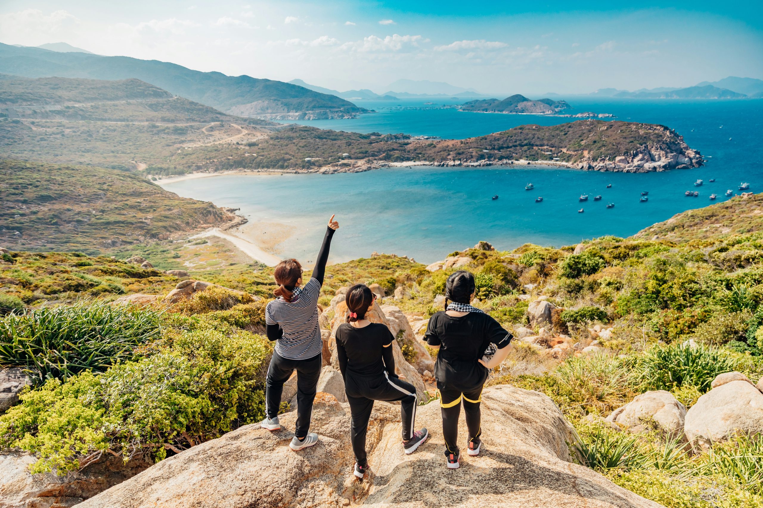 Drie wandelaars op een rotsachtig uitzichtpunt kijken uit over een tropische baai met boten en bergen.